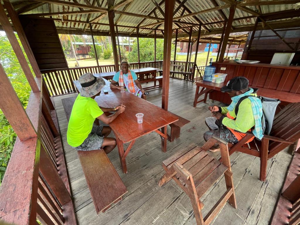 Drie personen zitten aan een houten tafel in een open loods, omringd door groen en natuurlijke elementen.